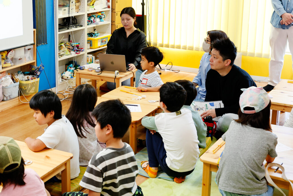 マシュマロチャレンジのイベント中、建物の仕組みやタワー作りのコツについて講義を受ける小学生と保護者たちの様子。デザイン思考を学び、次の挑戦に向けて意欲を高める場面。