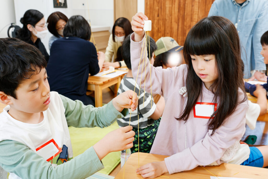 マシュマロチャレンジで、完成したタワーの頂上にマシュマロを慎重に乗せる小学生たちの様子。チームで協力しながら挑戦する、府中市開催の体験型ワークショップの一場面。