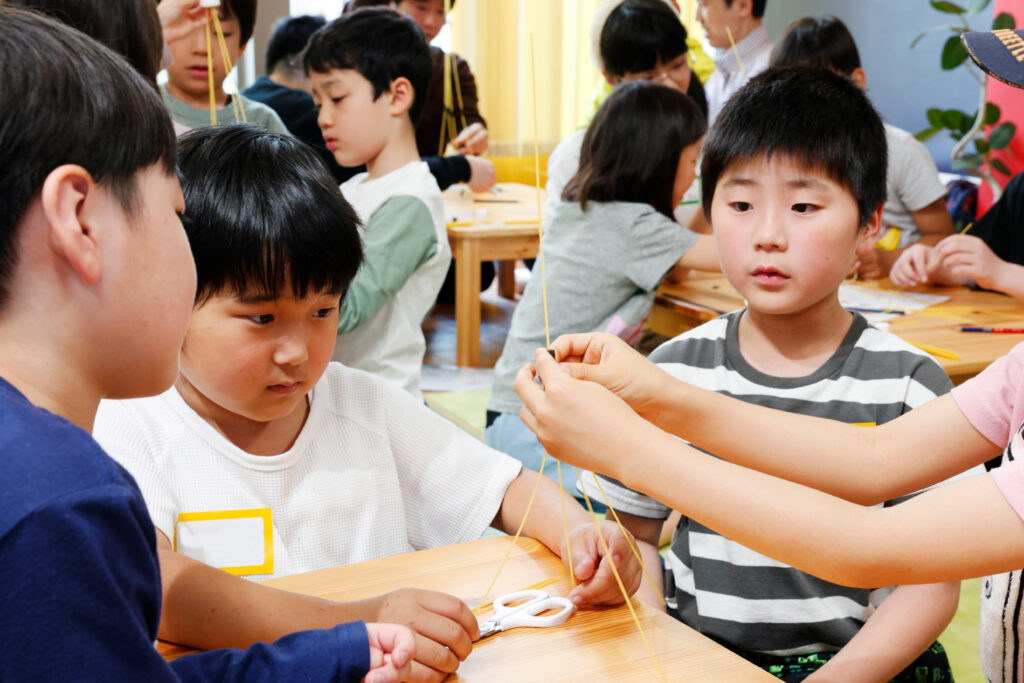 マシュマロチャレンジのタワー制作に挑戦する小学生たちの様子。スパゲッティを使ってチームで協力しながら、高い構造物づくりに集中しているシーン。