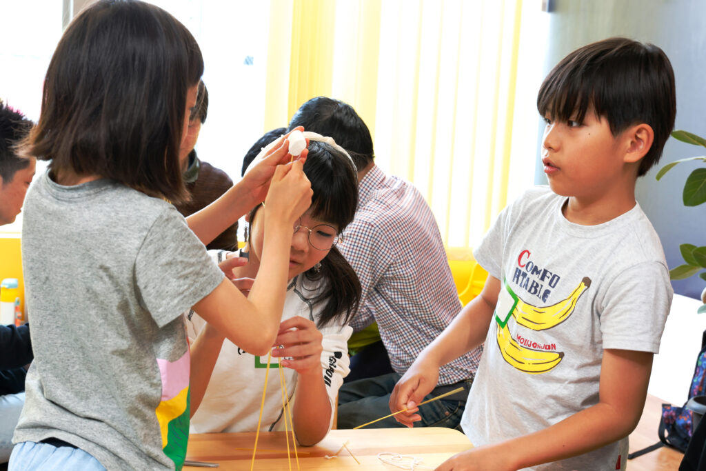 マシュマロチャレンジで、完成したパスタタワーの頂上にマシュマロを慎重に取り付ける小学生たちの様子。協力しながら最後の仕上げに挑む、創造力とチームワークが光る場面。