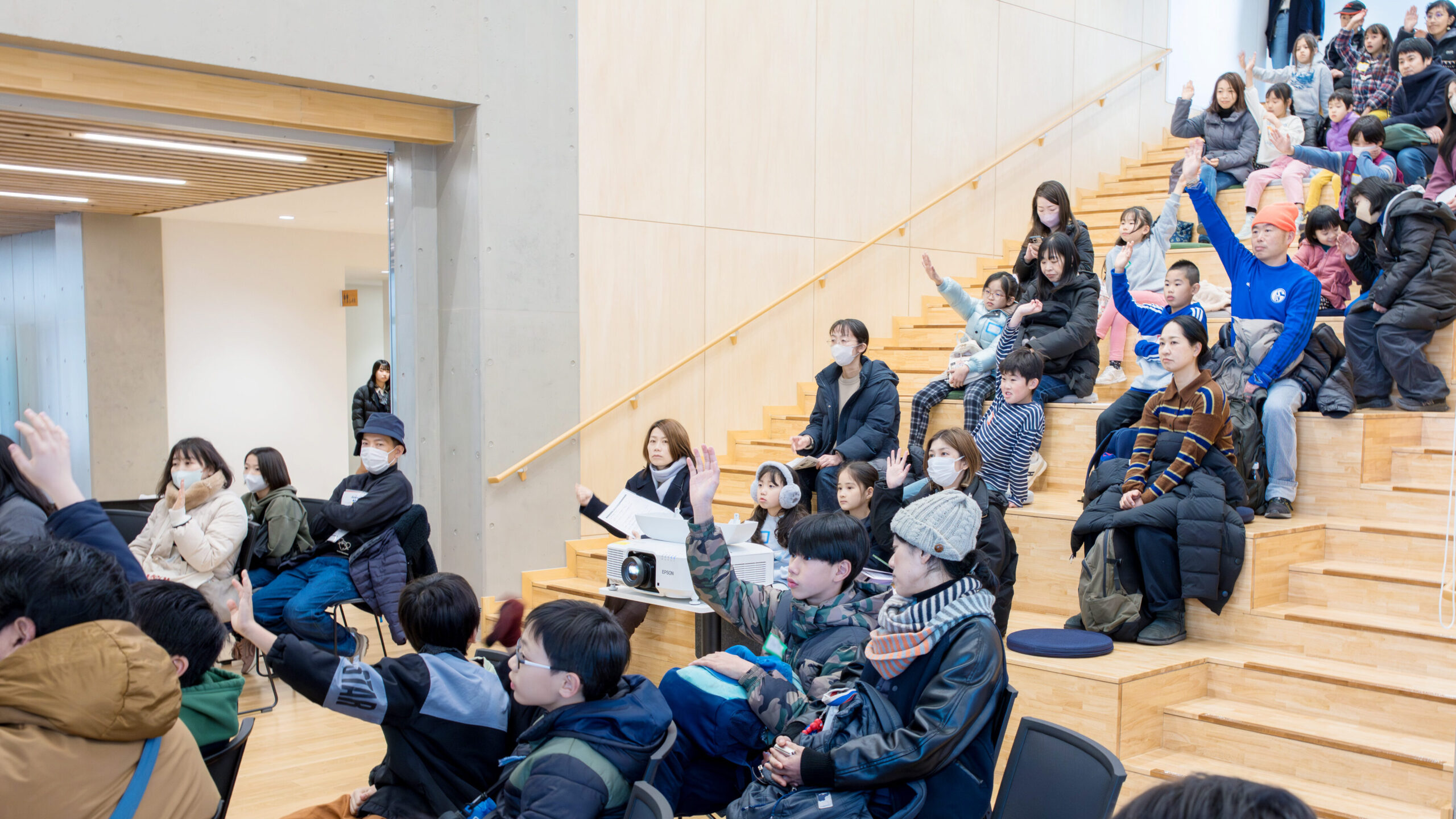 クイズ大会の様子。小学生と保護者が手を挙げて参加し、会場が盛り上がっているワンシーン。