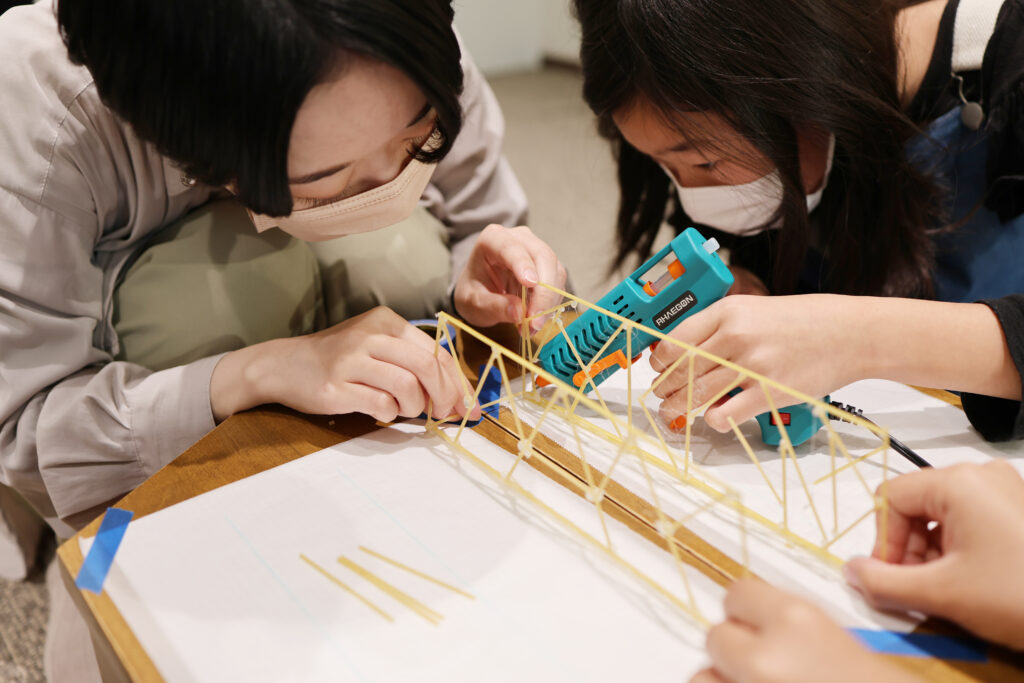 乾燥パスタで組んだトラス構造の橋を、グルーガンを使って組み立てる参加者たち