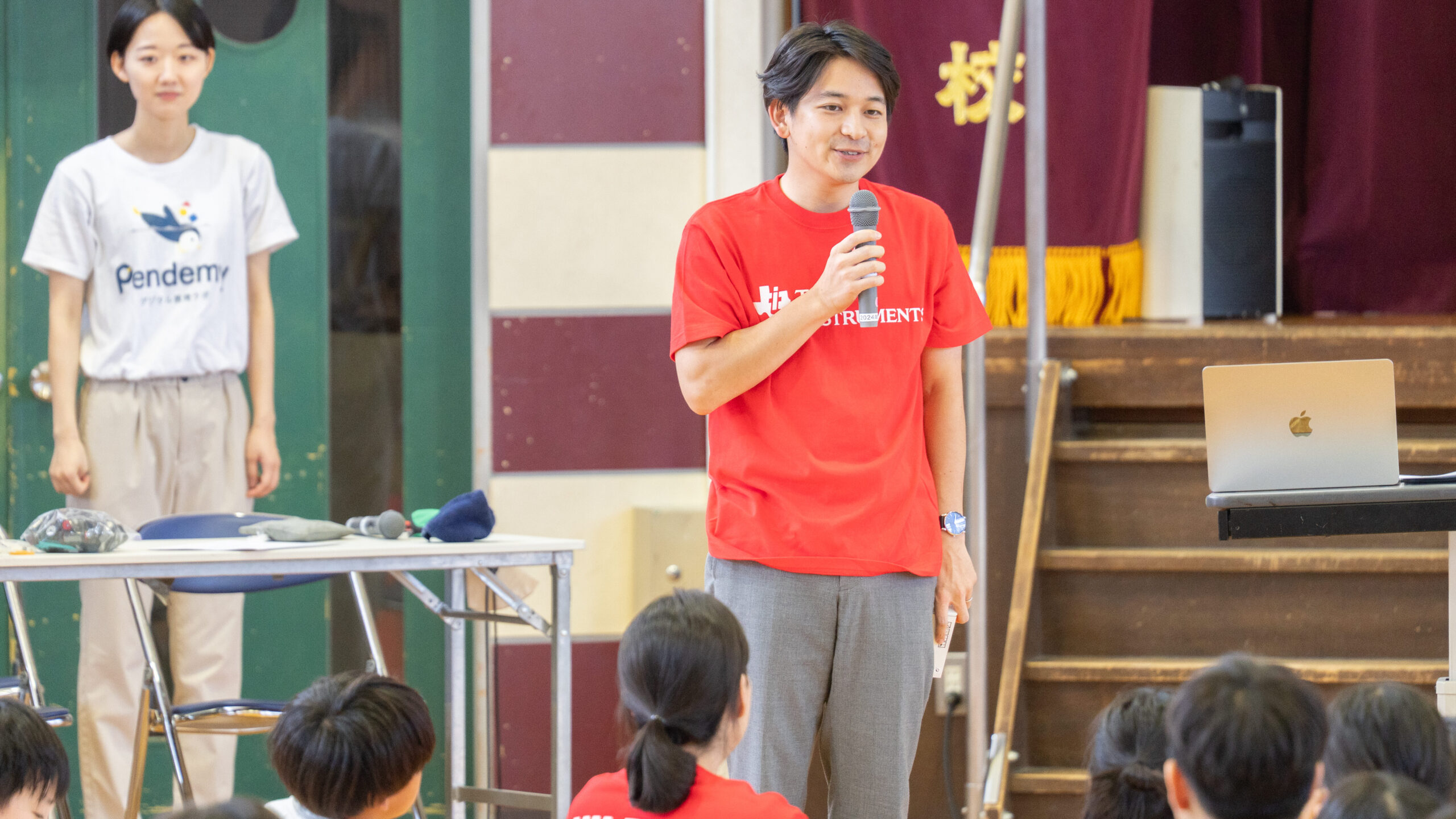 体育館のステージ前でマイクを持ちイベントの趣旨を説明するTI社員とPendemyスタッフ
