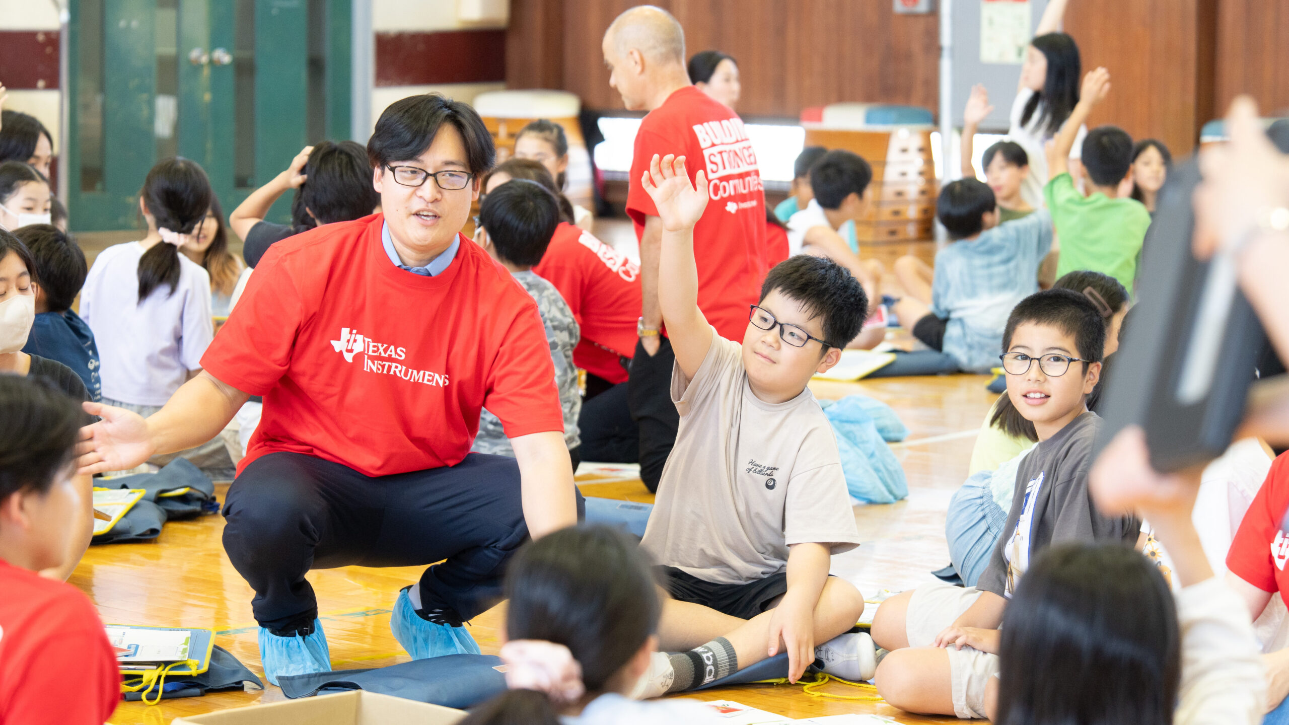 体育館の床に座り、問いかけに勢いよく手を挙げる小学生と、そばで見守るテキサス・インスツルメンツ社員