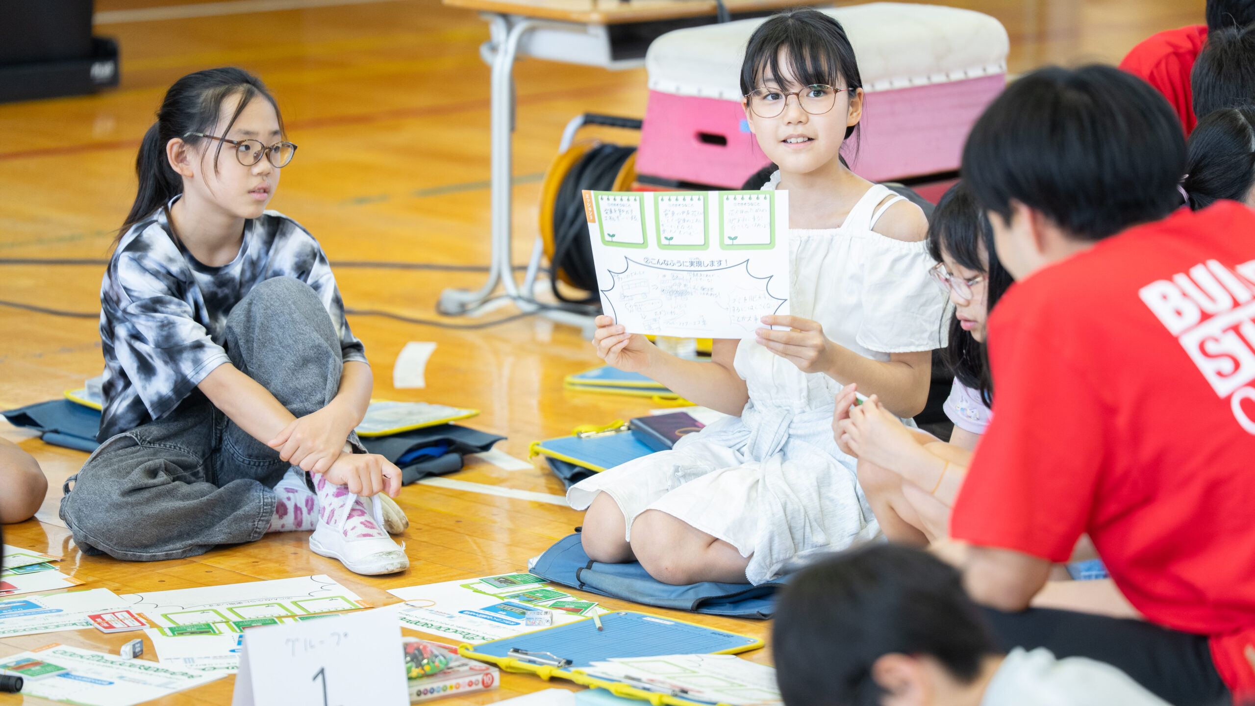 ワークシートを掲げて、自分が考えた施設のアイデアをグループのメンバーに説明する小学生