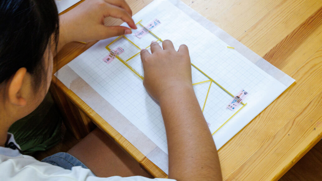 方眼の台紙の上にパスタを並べてマスキングテープで固定する子どもの手元