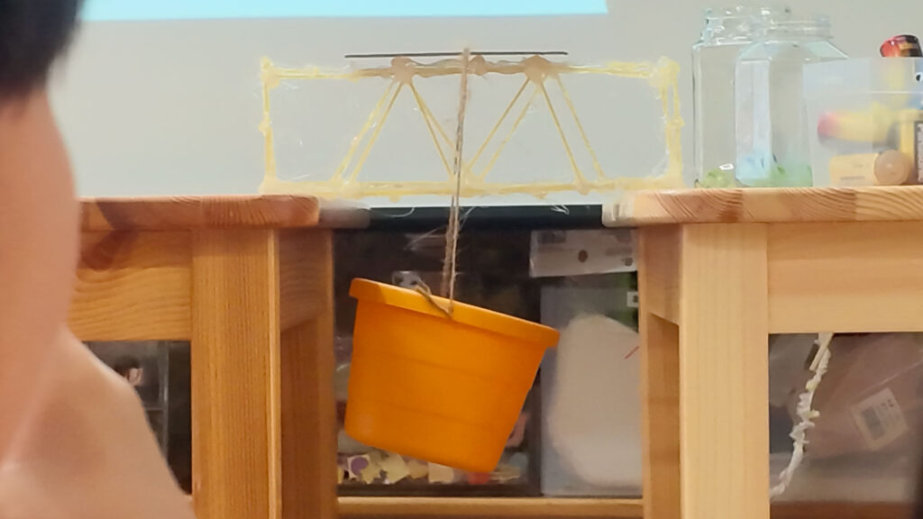 2台の机の間に渡したパスタ橋の中央から、電池を入れるオレンジ色のバケツをひもで吊るして強度を計測する様子